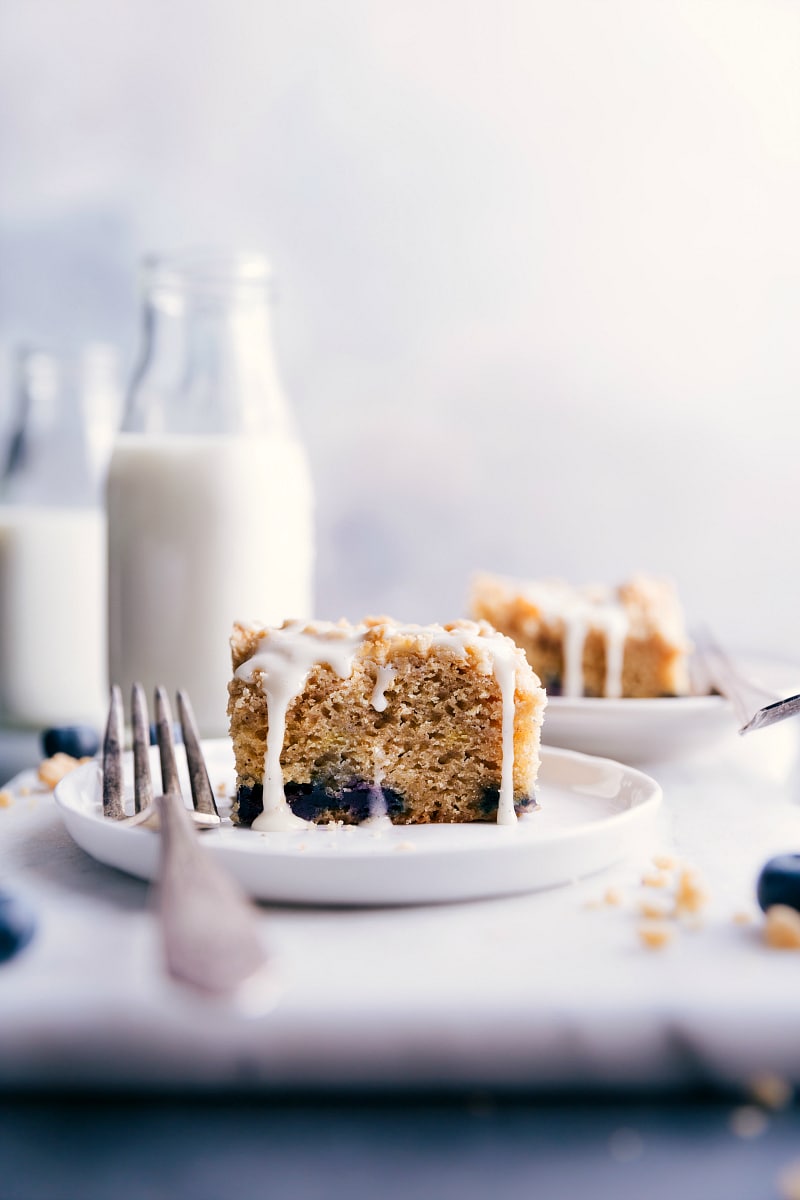 This moist and flavorful Blueberry Coffee Cake with a hint of lemon is topped with a sweet streusel and simple lemon glaze. Recipe via chelseasmessyapron.com #easy #lemon #buttermilk #withcrumbtopping #recipes #recipeseasy #best #blueberry #coffee #cake Image of a piece of coffee cake on a plate, with a fork on the side.