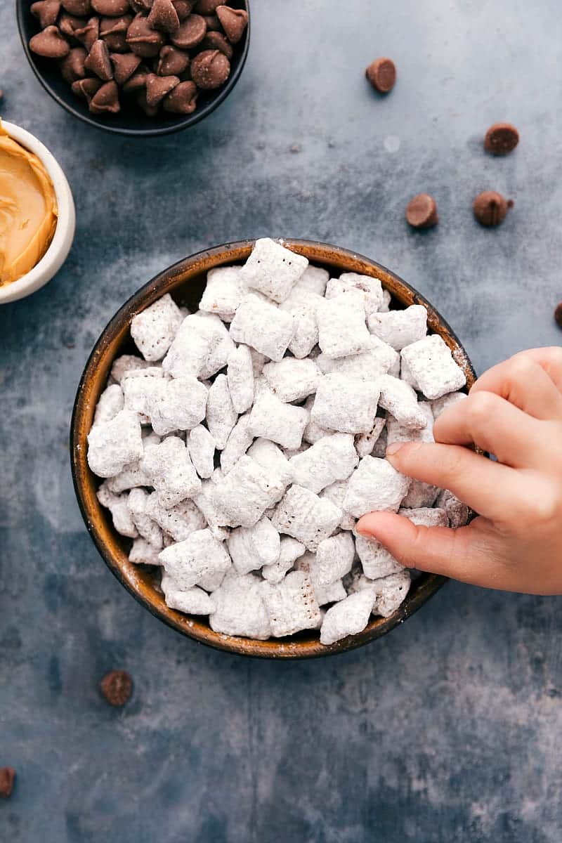 These Muddy Buddies (also called Puppy Chow) are the best easy treat. There's no baking required and only aย few ingredients!ย Recipe via ChelseasMessyApron.com #muddybuddies #puppychow #chex #chocolate #easy #treat #favorite Overhead photo of a bowl filled with Muddy Buddies and a toddler hand grabbing one out.