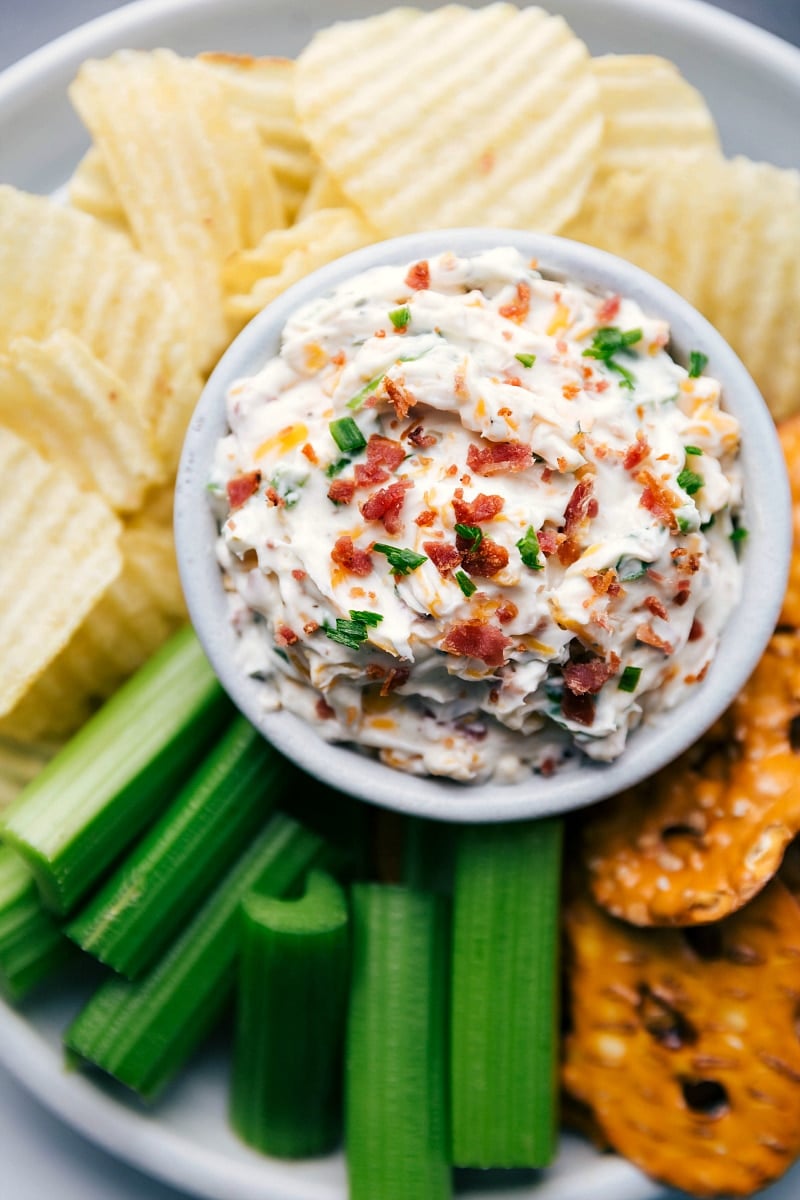 Baked Potato Dip Creamy and Topped with Herbs and Bacon Surrounded by Potato Chips and Celery Sticks Baked potato dip creamy and topped with herbs and bacon surrounded by potato chips and celery sticks.