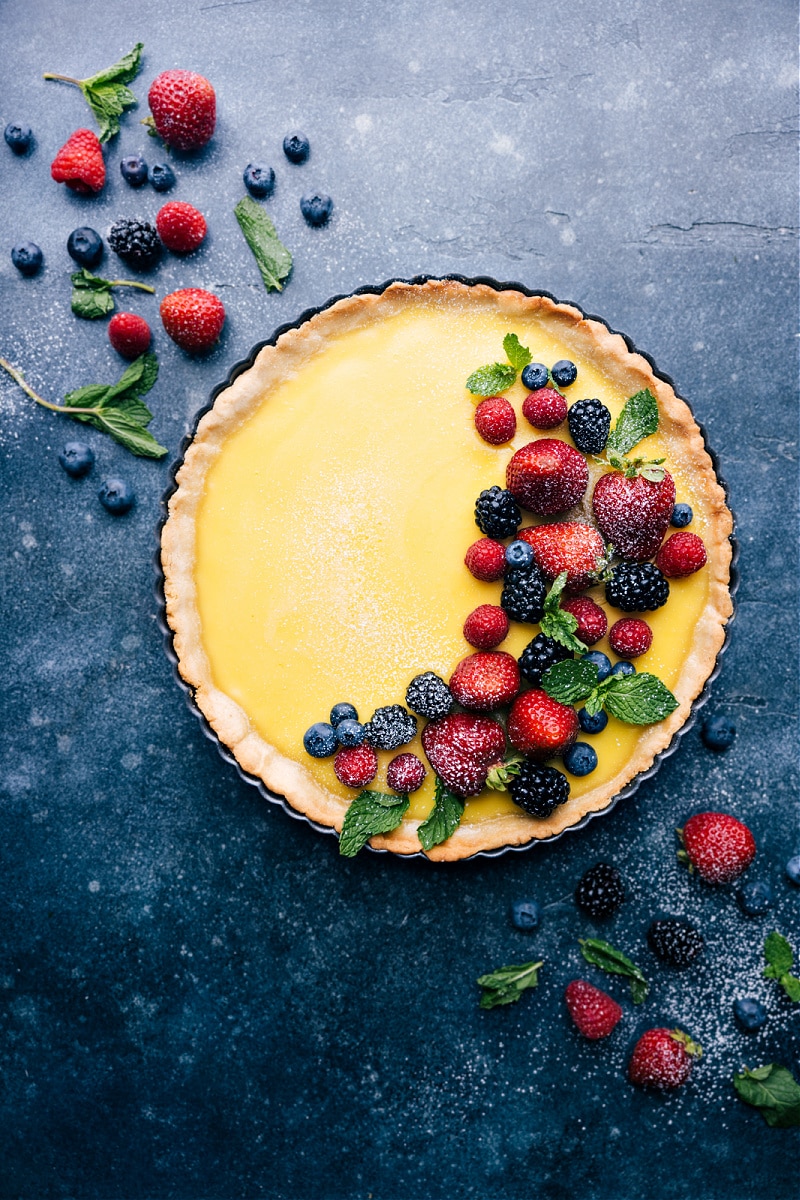 This Lemon Tart starts with a thick, sweet, and buttery crust which we load with a creamy, custardy tart lemon filling. Top your tart with fresh berries and a sprinkle of powdered sugar for the perfect finishing touch! Recipe via ChelseasMessyApron.com #lemontart #betterthanpie #custart #tartsweet #lemony #dessert Overhead view of the Lemon Tart