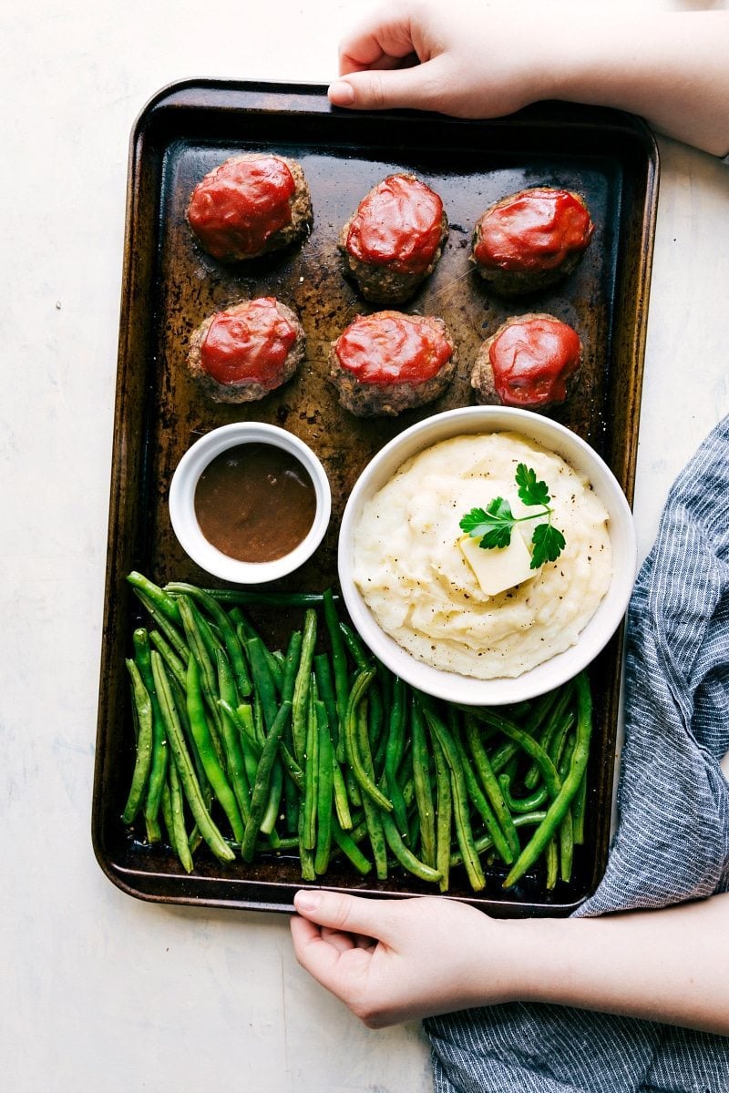 Mini Meatloaf, mashed potatoes, and roasted green beans all get baked together on ONE sheet pan! Recipe via chelseasmessyapron #easy #meat #sheetpan #recipe Overhead shot of the mini meatloaves all on one pan with potatoes, green beans, and gravy