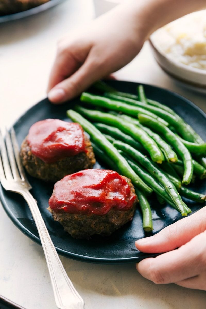 Best mini meatloaf recipe, mashed potatoes, and roasted green beans all get baked together on ONE sheet pan! Recipe via chelseasmessyapron #easy #meat #sheetpan #recipe image of two mini meatloafs on a plate with green beans and a fork on the side