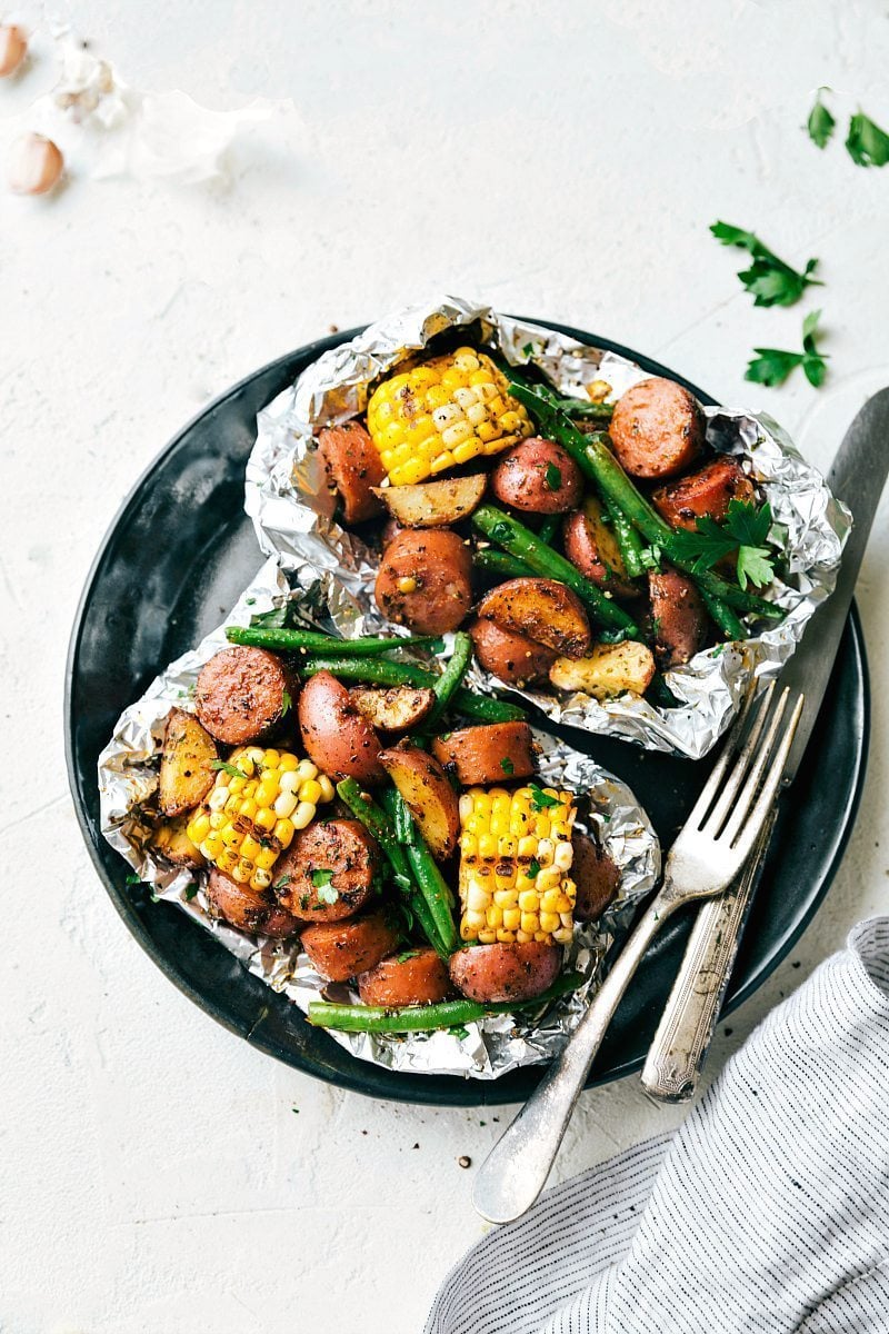 Easy Foil Pack Garlic Butter Sausage & Veggies with smoked sausage, red potatoes, sweet corn, and crisp green beans. Toss everything with a simple (but flavor-packed) seasoning blend, add some butter, and throw these packets on the grill or in the oven. These simple foil pack dinners can be prepped out in 15 minutes or less! Recipe via ChelseasMessyApron #dinner #quick #easy #simple #camping #overfire #grilled #garlicbutter #sausage #veggies #foilpacks Overhead image of two garlic butter sausage and veggies in foil packs