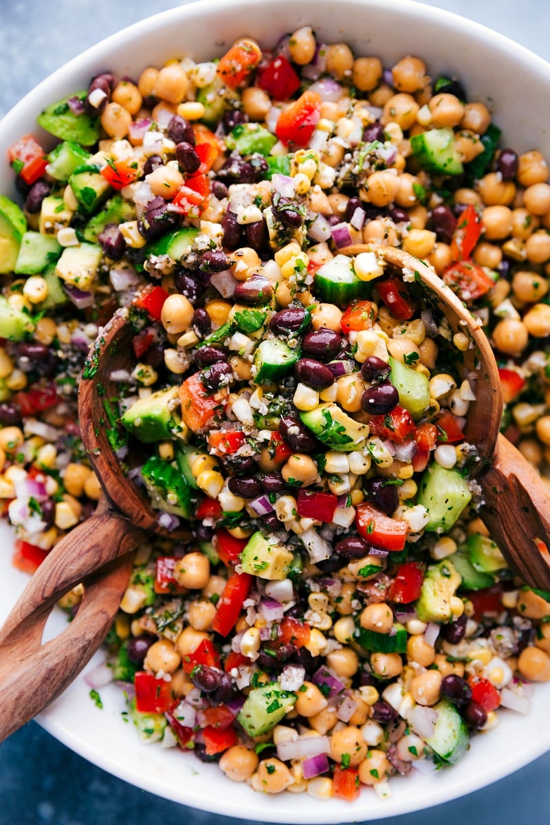 Garbanzo Bean Salad is loaded with veggies, dressed in an Italian-inspired vinaigrette, and finished with some herbaceous parsley! Recipe via chelseasmessyapron #protein #chickpeas #blackbeans #corn #avocado #redpepper #onion #cucumber #whole #healthy #best #quick #easy #simple #garbanzo #bean #salad #vegetarian Overhead image of Garbanzo Bean Salad in a bowl with serving spoons
