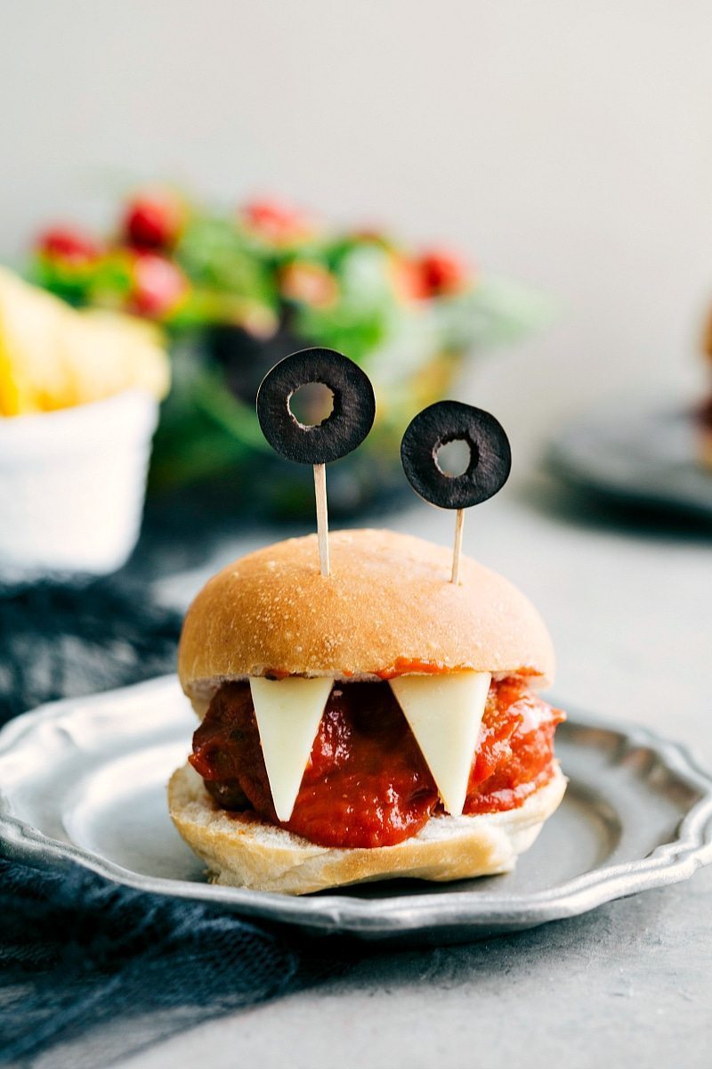 Monster Meatball Sliders: Four easy, & creative Halloween appetizers that are so delicious -- Spiderweb 7-Layer Dip, Walking Dead Mummy Dogs, Monster Meatball Sliders, & Cheesy Monster Fingernail Bread. chelseasmessyapron.com Image of the Monster Meatball Sliders with olive eyes and cheese teeth that go along with these halloween appetizers easy to make