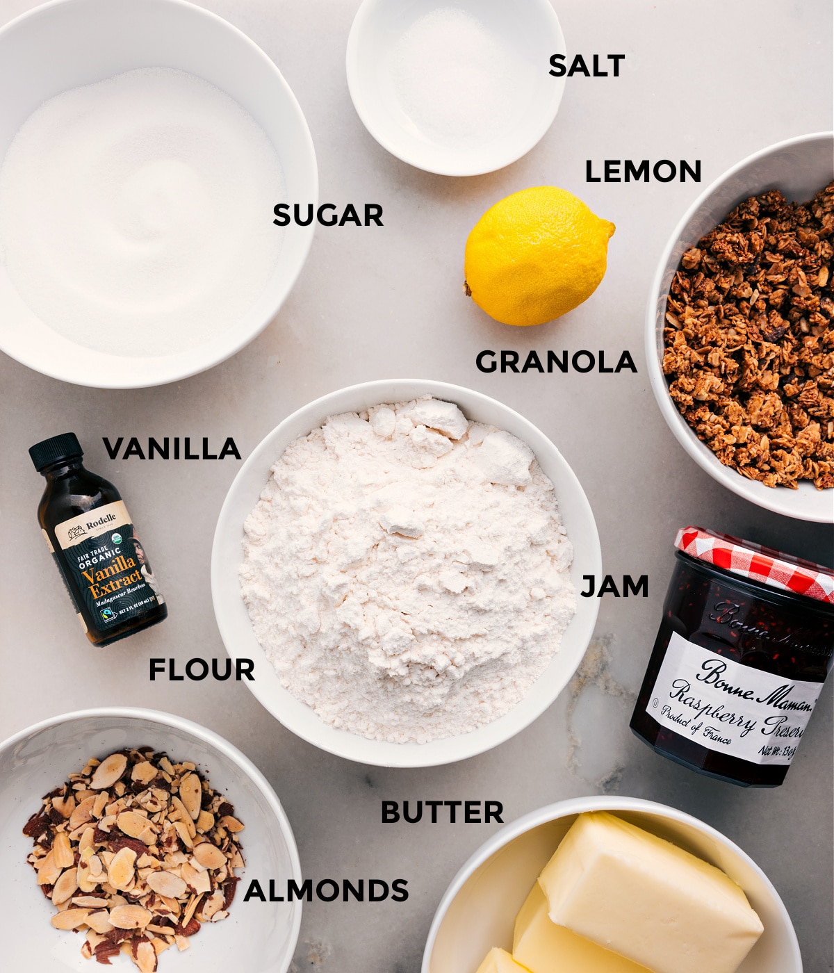 Ingredients In Raspberry Bars All the ingredients prepped and ready for easy assemblyโlemon, granola, jam, flour, vanilla, sugar, salt, butter, and almonds.