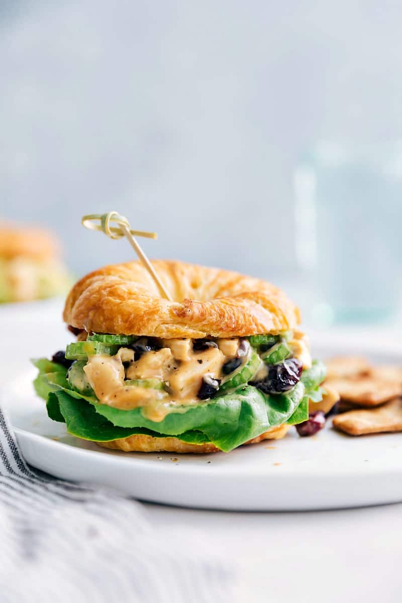 These delicious (and easy) Honey Mustard Chicken Salad Sandwiches are a fun twist on typical chicken salad! They're a hit wherever they're served! via chelseasmessyapron.com #honey #mustard #chicken #salad #easy #quick #lunch #dinner #party #holiday #potluck #lettuce #sandwich #croissant #celery #cranberries #rotisserie #best Close-up view of a Honey Mustard Chicken Salad Sandwich with crackers on the plate and water in the background.