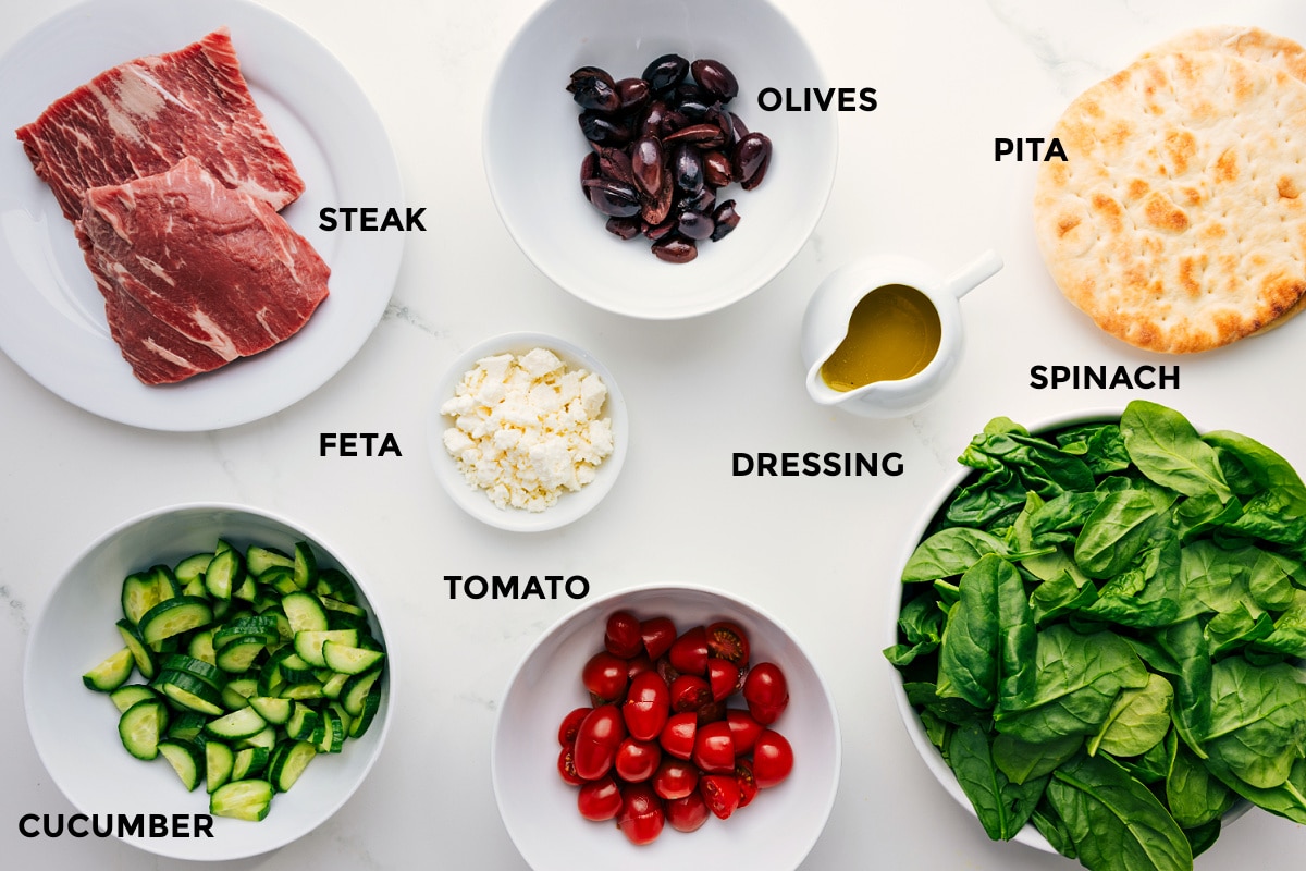 Ingredients In Grilled Steak Salad Recipe All the ingredients prepped for easy assembly, including sliced steak, olives, pita wedges, spinach, honey-lemon dressing, crumbled feta, cherry tomatoes, and cucumber.