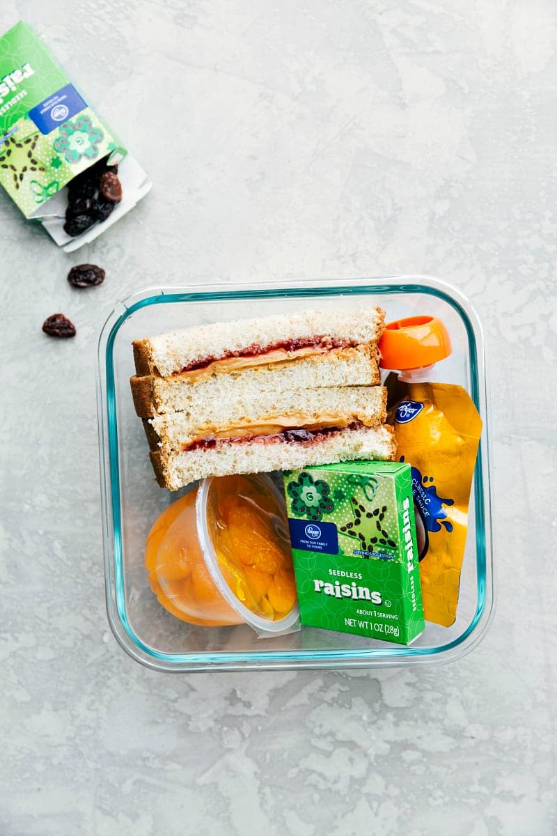 Packed School Lunch with Sandwich and Fruits A container filled with a peanut butter and jam sandwich, raisins, mandarin oranges, and apple sauce, all neatly arranged and ready to go.