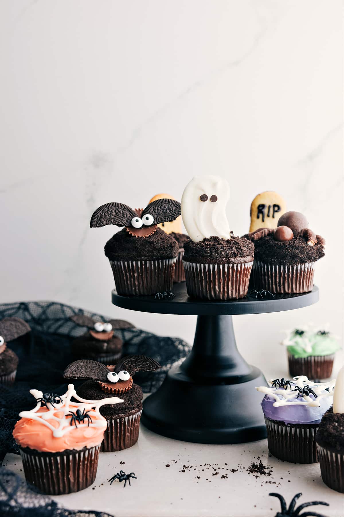 Halloween cupcakes displayed on a cupcake holder feature a ghost, spider, bat, and tombstone cupcakes.