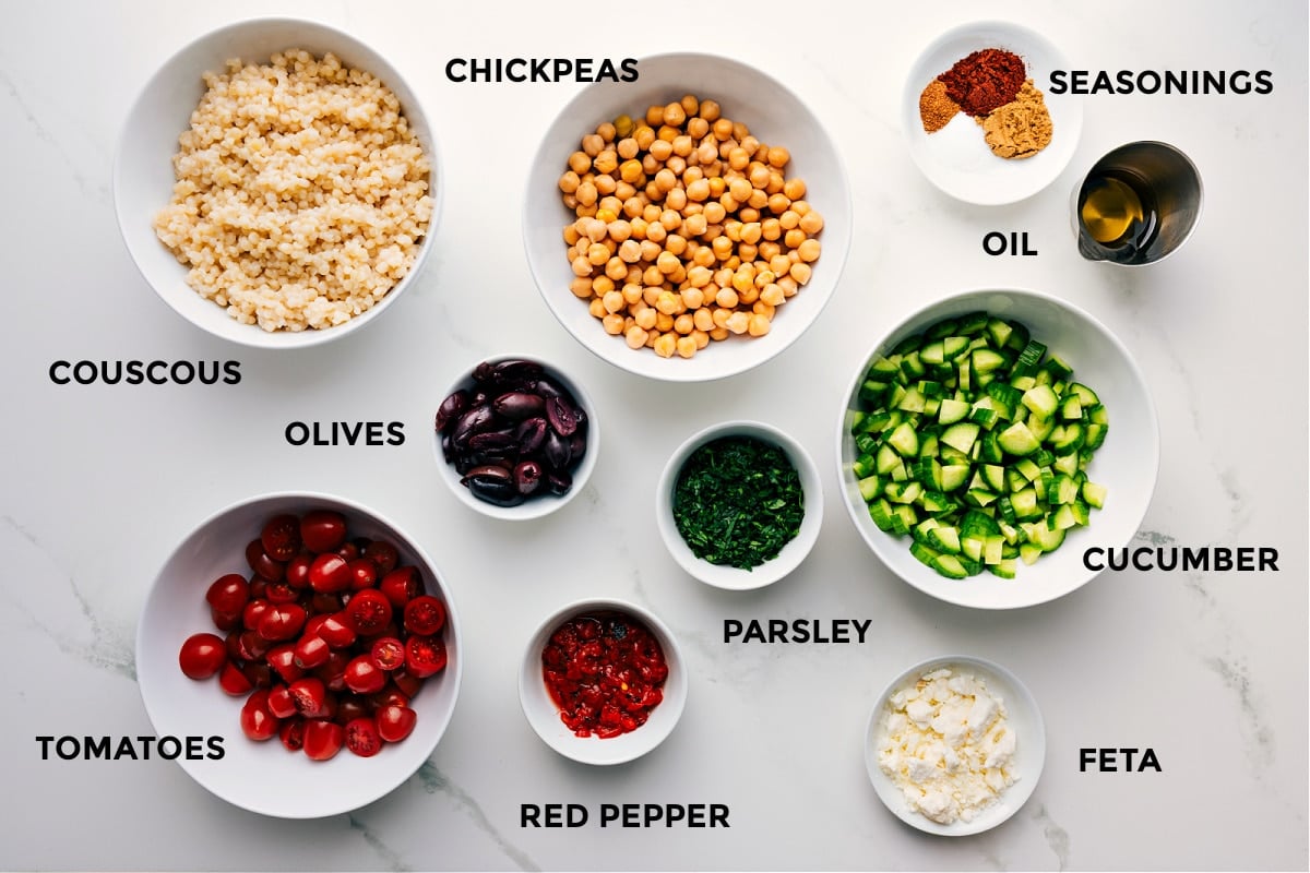 Ingredients In Chickpea Salad All the ingredients prepped for easy assembly, including chickpeas, couscous, seasonings, oil, cucumbers, parsley, feta, peppers, tomatoes, and olives.
