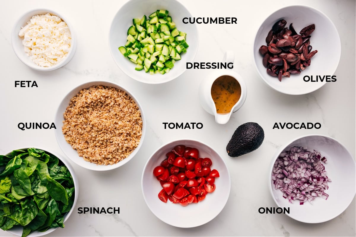 Ingredients In Greek Quinoa Salad Ingredients in this recipe prepped out for easy assembly including the feta, quinoa, spinach, cucumber, dressing, olive, avocado, onion, and tomato.