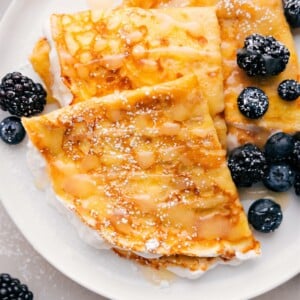 Plate of three crepes with berries and syrup drizzled on top.