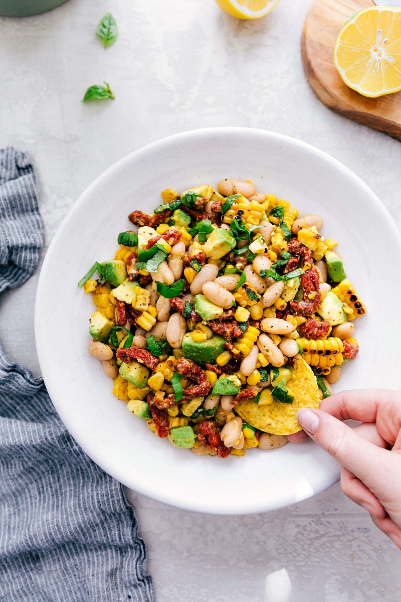 White Bean Salsa! Recipe via Chelseasmessyapron #healthy #keto #whole #protein #whole30 #best #avocado #corn #sundriedtomatoes #clean #cleaneating #whitebean #salsa Overhead image of the White Bean Salsa with a chip being dipped