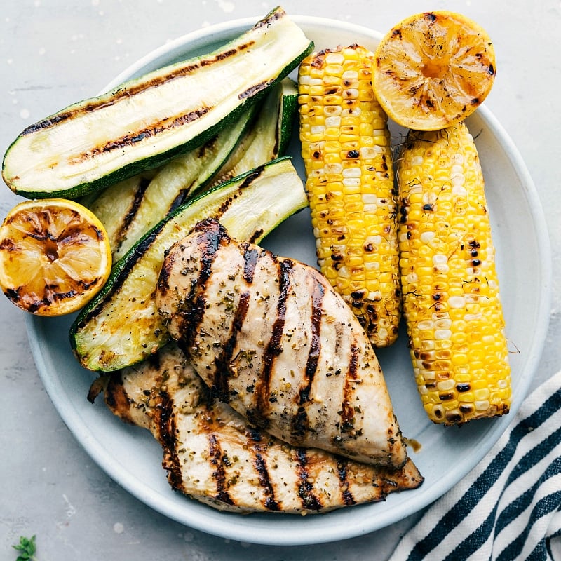 Greek Chicken Recipe The result of perfectly marinated and grilled meat, served with grilled zucchini, corn, and lemons on the side, creating a mouthwatering meal.