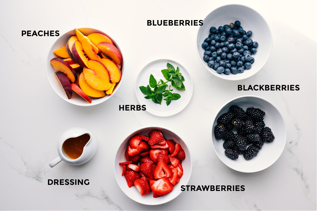 Ingredients In Peach Fruit Salad Ingredients in this recipe prepped out for easy assembly including peaches, herbs, blueberries, blackberries, strawberries, and dressing.