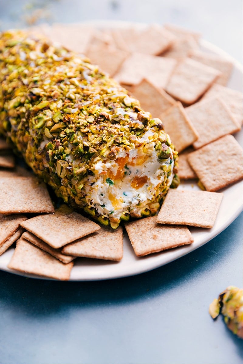 This delicious Pistachio Goat Cheese Ball is one for the books! Tangy goat cheese paired with sweet dried fruit, fresh herbs, and encased with salty pistachios -- your next party needs this appetizer! Recipe via ChelseasMessyApron #appetizer #gameday #snack #best #quick #easy #crackers #familyfriendly #pistachio #goatcheese #ball Up-close image of the pistachio goat cheese ball on a plate with crackers on the side ready to be enjoyed