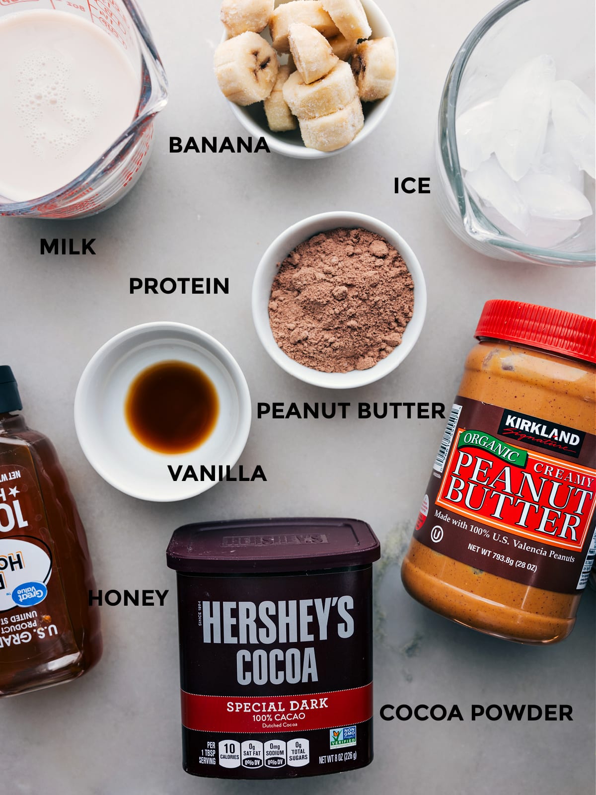 Ingredients In Peanut Butter Protein Shake Recipe All the ingredients including cocoa powder, vanilla, peanut butter, protein powder, bananas, milk, ice, and honey prepped out for easy assembly.