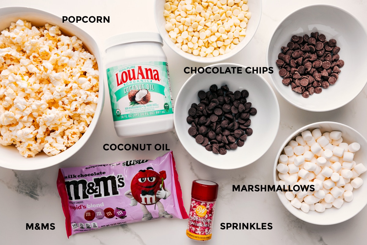 Ingredients In Valentine's Popcorn All the ingredients in this recipe prepped out for easy assembly including the popcorn, coconut oil, chocolate chips, marshmallows, M&Ms, and sprinkles.