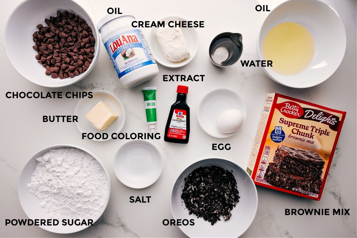 Ingredients In Mint Brownies Recipe All the ingredients prepped and ready for easy assembly, including chocolate chips, butter, food coloring, powdered sugar, salt, oil, cream cheese, extract, Oreos, water, egg, and brownie mix.