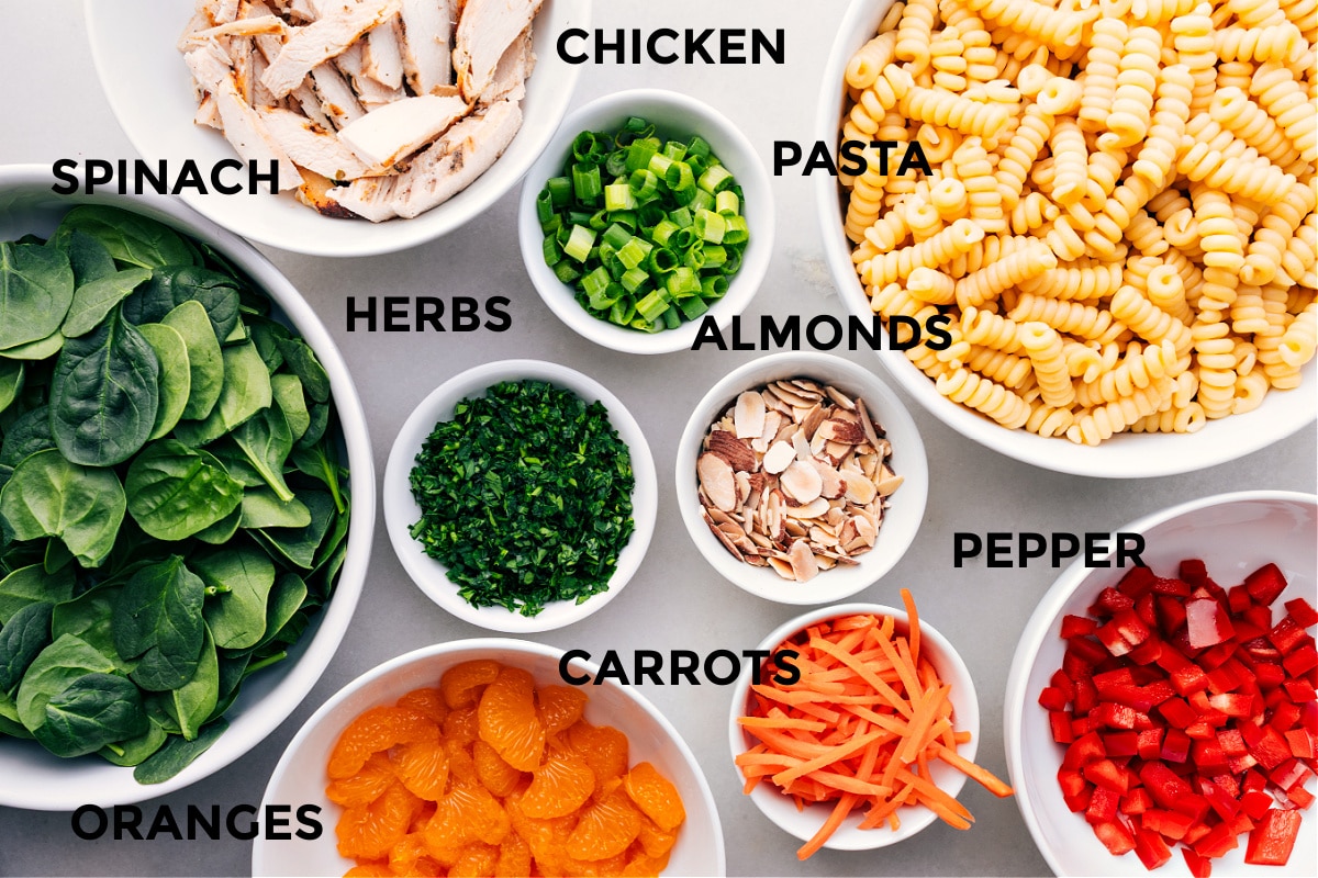 Ingredients In Asian Pasta Salad Asian pasta salad ingredients laid out for easy assembly including chicken, pasta, almonds, herbs, spinach, peppers, carrots, and oranges.