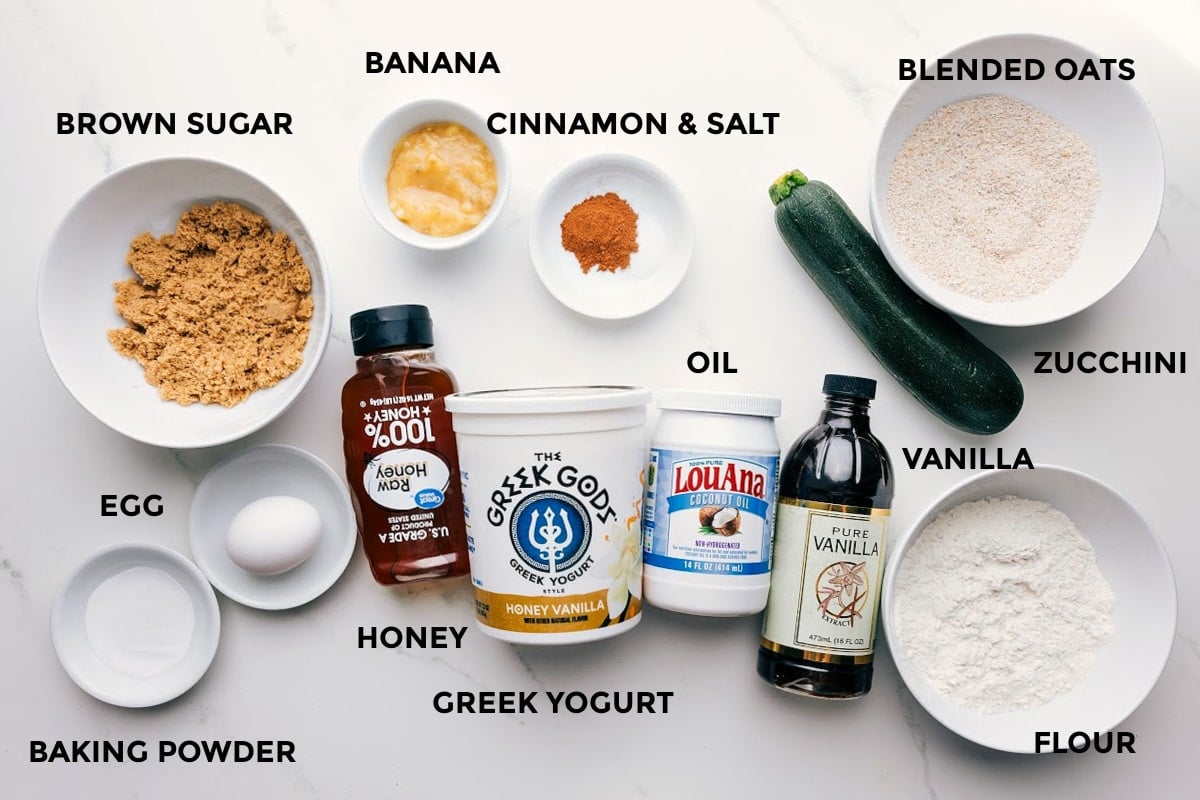 All the ingredients in this recipe prepped out for easy assembly including the brown sugar, egg, baking powder, honey, Greek yogurt, banana, cinnamon, salt, oil, vanilla, flour, zucchini, and blended oats.