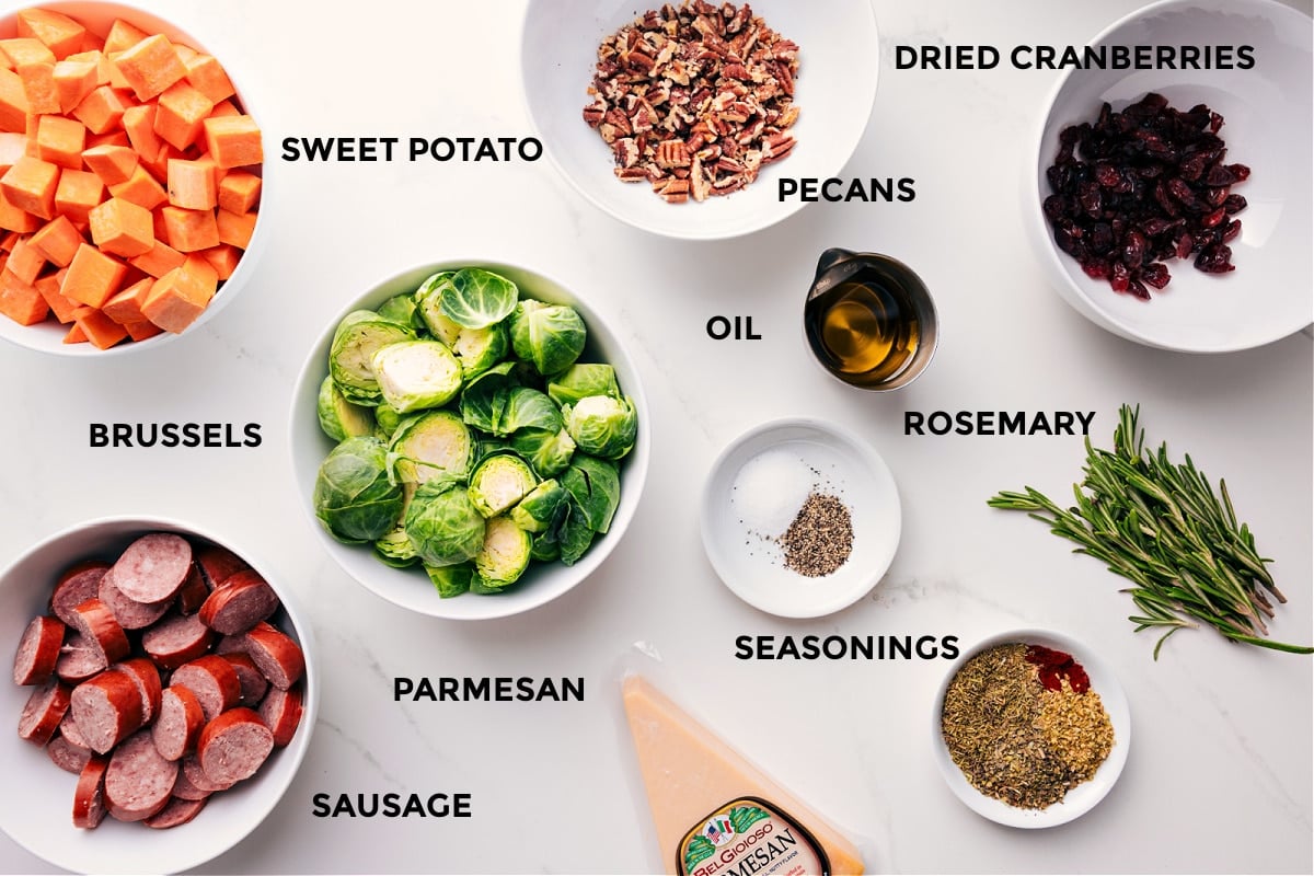 Ingredients In Prepped ingredients for sheet pan: sweet potatoes, sausage, Brussels sprouts, rosemary, cranberries, pecans, Parmesan.