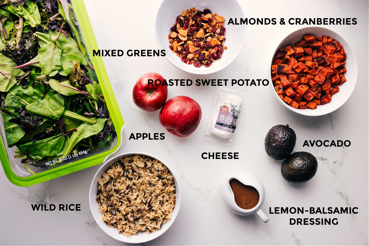 Hearty and nutritious Roasted Sweet Potato Salad. All the ingredients prepped out for easy assembly including mixed greens, wild rice, almonds, cranberries, roasted sweet potatoes, avocado, cheese, and dressing.