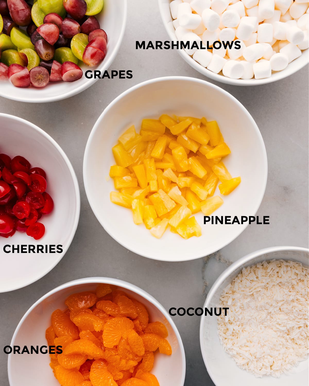 Ingredients Prepped Out For Ambrosia Salad The ingredients in this recipe include pineapple, marshmallows, grapes, cherries, coconut, and oranges.