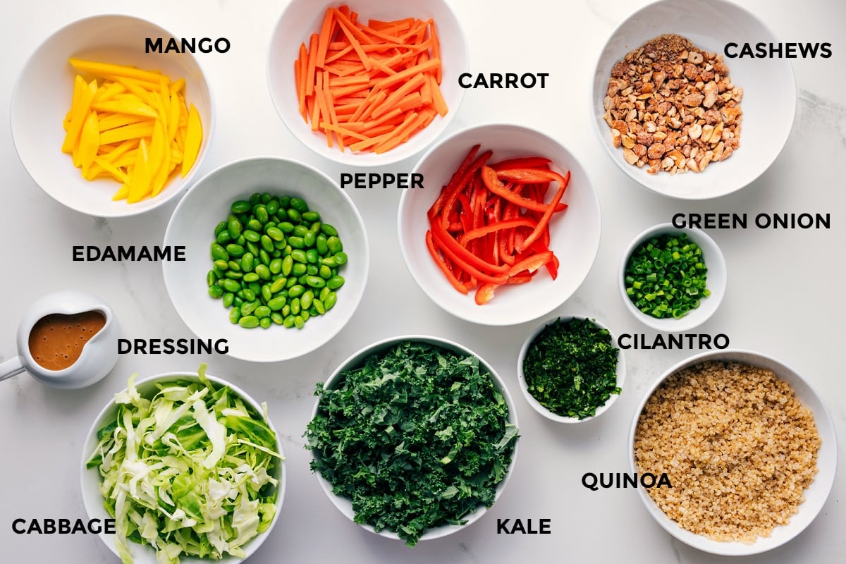 Ingredients In This Thai Quinoa Salad All the ingredients in this recipe prepped out for easy assembly including the mango, dressing, cabbage, edamame, pepper, carrot, kale, cilantro, green onion, quinoa, and cashews.