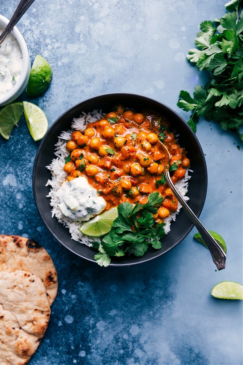 This flavor-packed Chickpea Curry is loaded with aromatic spiced veggies, tender chickpeas, and a creamy coconut sauce. This plant-based curry is an easy weeknight meal that is naturally gluten-free and vegan. Recipe via ChelseasMessyApron.com #chickpeas #curry #garbanzo #coconut #raita #vegan #vegetarian #naan Chickpea Curry, served with rice, cilantro, naan bread and coconut raita.