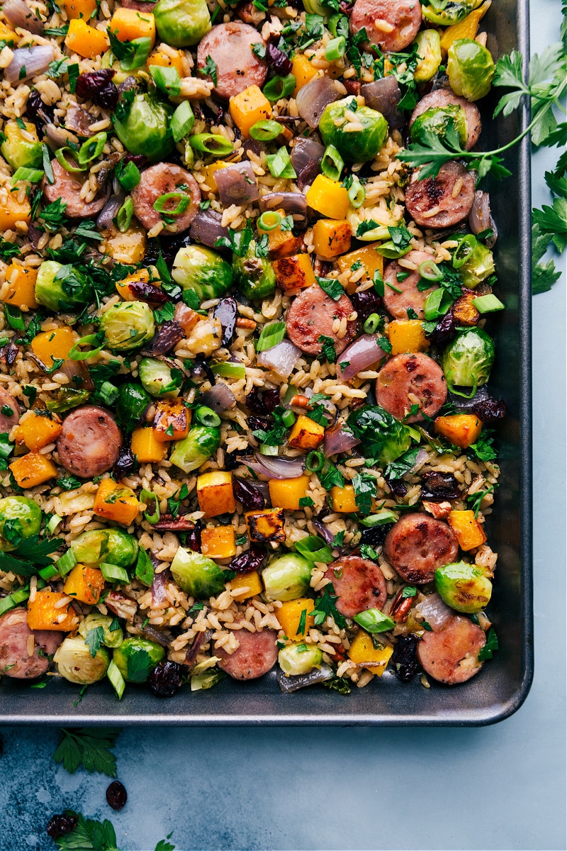 Butternut Squash Wild Rice Pilaf is a tasty and unique ONE-PAN meal! We roast together seasoned wild rice, smoked chicken & apple sausage, maple butternut squash, and perfectly caramelized brussels sprouts and then drizzled in an irresistible maple-orange vinaigrette. (That's right -- even the wild rice cooks on the sheet pan along with everything else!) Recipe via ChelseasMessyApron #dinner #quick #easy #simple #best #butternutsquash #wildrice #pilaf Overhead view of Butternut Squash Wild Rice Pilaf in the pan