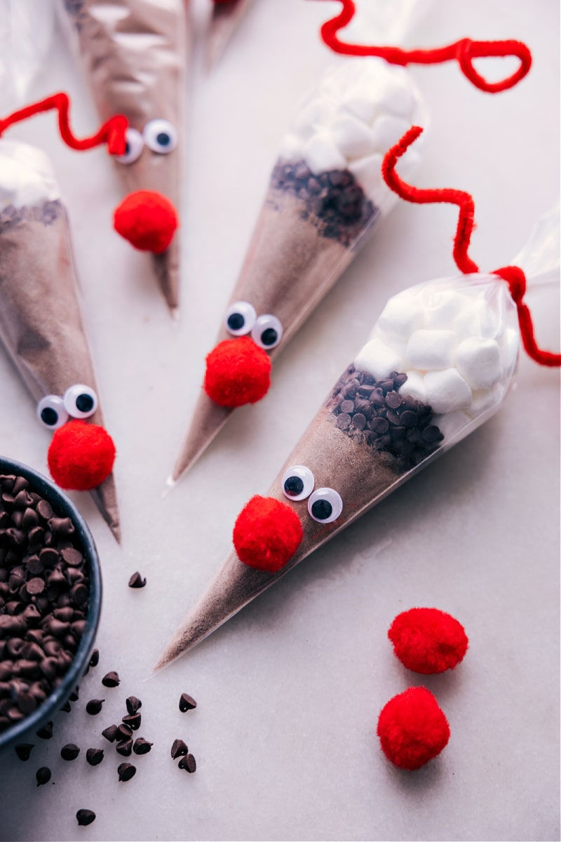 The cutest edible gift this holiday season! Add hot chocolate mix, chocolate chips, and marshmallows to a cone-shaped bag and decorate to look like Reindeer (specifically Rudolph). Put a smile on your kid's face or gift out to neighbors, friends, co-workers, family, etc. Recipe via chelseasmessyapron #dessert #gift #christmas #holidays #christmasgifts #holidaygifts #rudolph #reindeer #hot #chocolate Overhead image of the Reindeer Hot Chocolate ready to be given out