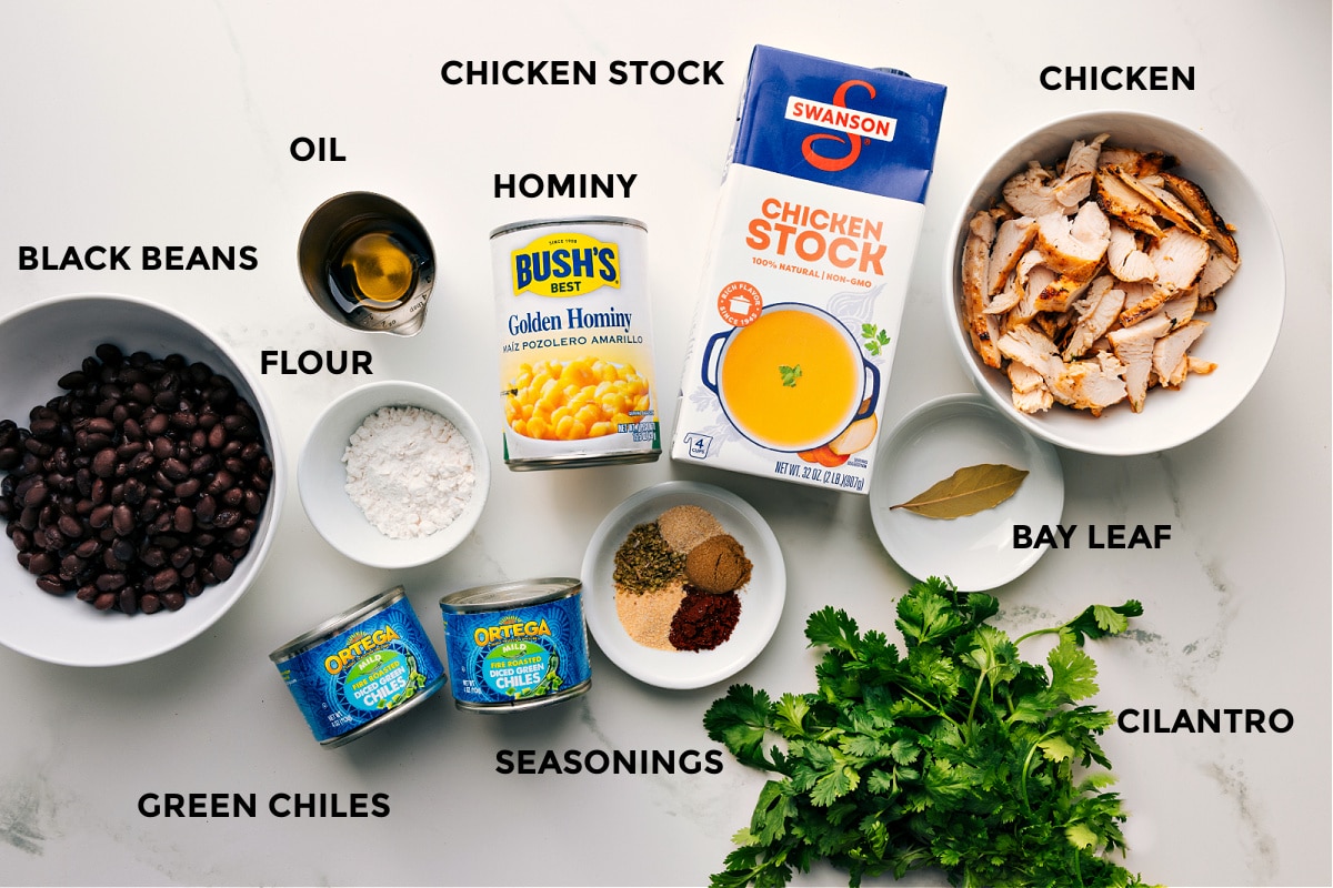 Ingredients In Chicken Pozole Prepped ingredients for this recipe laid out for easy assembly, including black beans, flour, green chiles, seasonings, oil, hominy, stock, bay leaf, chicken, and cilantro.