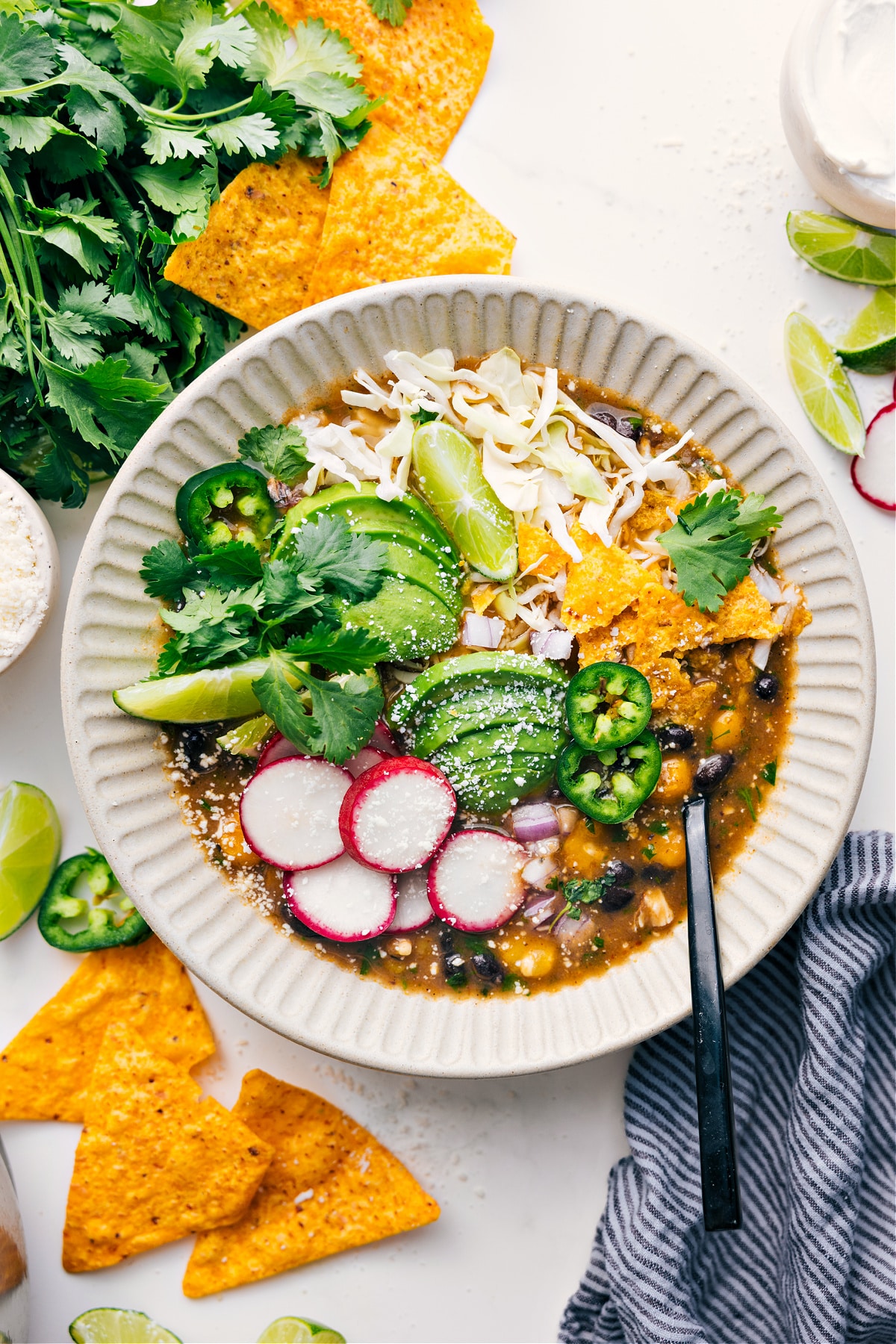 Chicken Pozole served in a bowl topped with shredded cabbage, tortilla chips, avocado slices, radishes, cilantro, lime wedges, and jalapeño.
