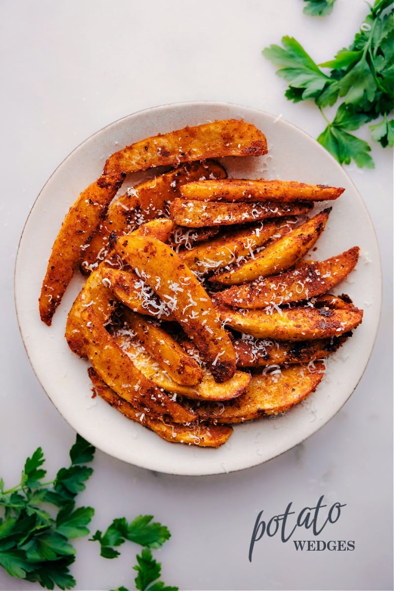 Looking for a healthier alternative to french fries that doesn't skimp on flavor and texture? Look no further than these crispy, fluffy baked Potato Wedges! These potatoes are the epitome of perfection, with a crisp exterior and soft, pillowy interior that is completely irresistible. Best of all, they're a budget-friendly and filling side dish that can be paired with just about anything. So skip the deep fryer and whip up a batch of these potatoes for your next meal! Recipe via chelseasmessyapron #sidedish #best #quick #easy #simple #comfortfood #fries #potato #wedges Crispy potato wedges served on a white plate.