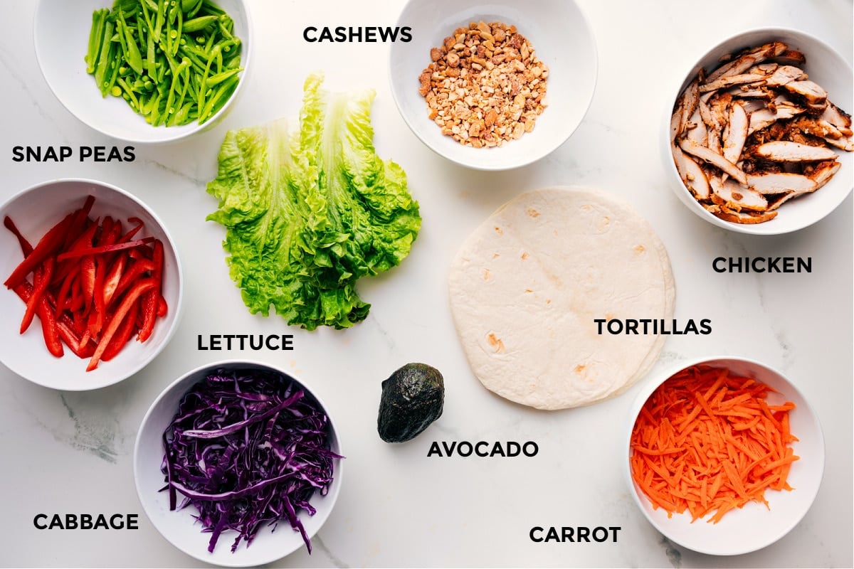 Ingredients in cashew chicken wraps prepped out for easy assembly including the snap peas, peppers, cashews, chicken, tortillas, carrot, avocado, lettuce, and cabbage.