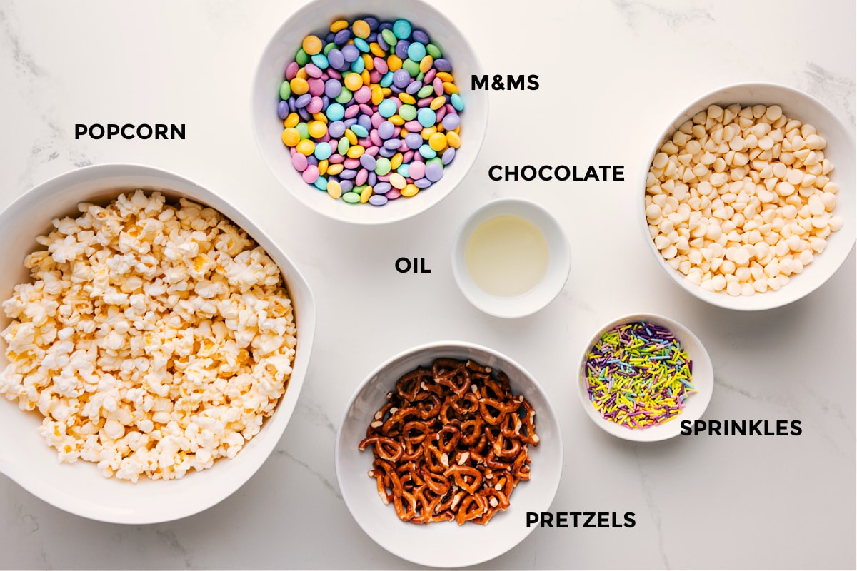 Ingredients In Easter Popcorn All the ingredients in this recipe prepped out for easy assembly including the popcorn, pretzels, M&Ms, sprinkles, chocolate, and oil.