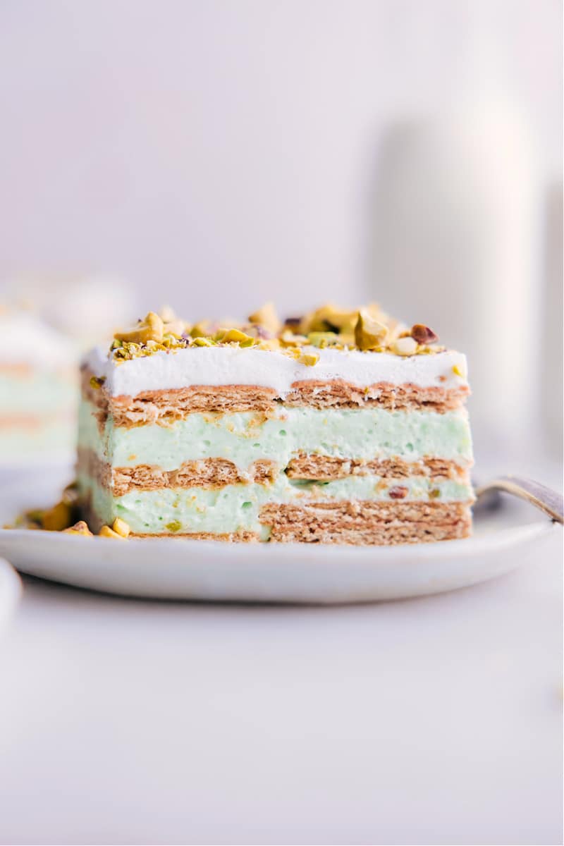 Pistachio Icebox Cake is made with layers of graham crackers and fluffy pistachio pudding, topped with whipped cream and crunchy pistachios, creating a perfect balance of sweet and nutty flavors with a creamy and smooth texture. This is an icebox cake, which means it's chilled in the refrigerator overnight, making it a convenient make-ahead dessert option. Recipe via chelseasmessyapron #dessert #best #quick #easy #simple #stpatricksday #treat #nobake #pistachio #icebox #cake Image of a slice of Pistachio Icebox Cake, ready to be enjoyed