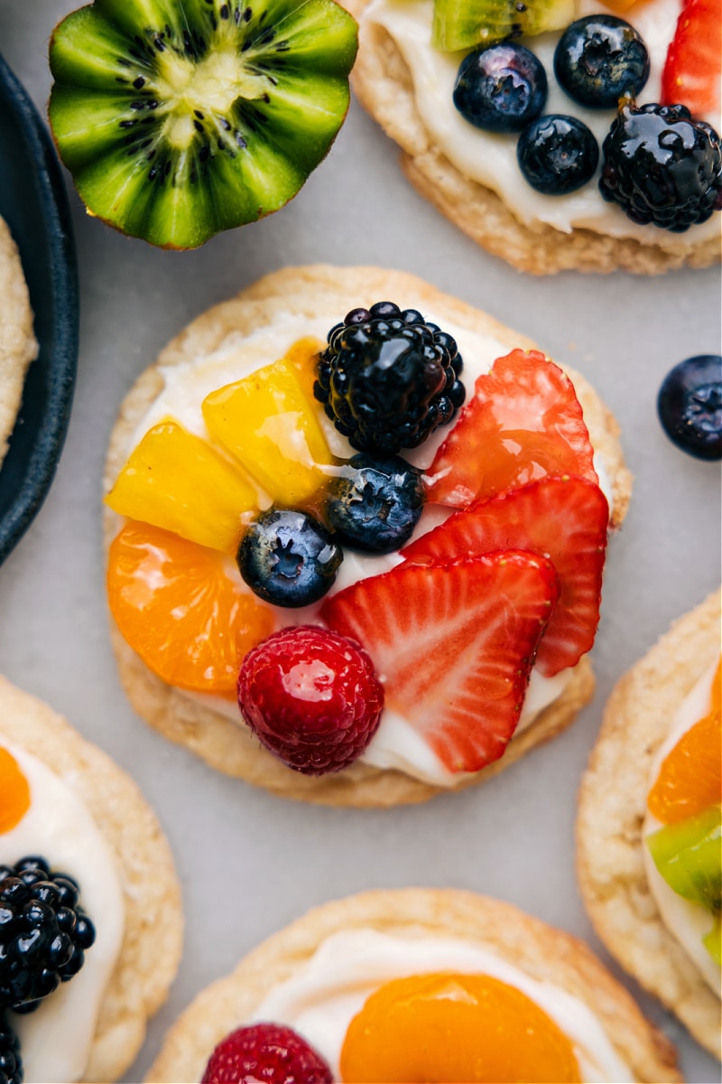 These Sugar Cookie Fruit Pizzas begin with soft, buttery sugar cookies that are topped with a velvety smooth and creamy frosting. Recipe via chelseasmessyapron #dessert #best #quick #easy #simple #fruity #sweettreat #sugarcookie #fruitpizzas Up close overhead image of the dessert ready to be enjoyed
