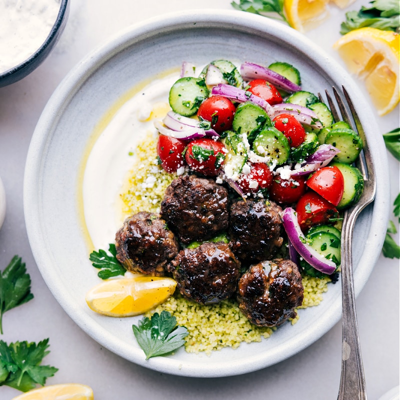 These insanely delicious Greek Meatball Bowls make for a satisfying meal! Featuring juicy meatballs seasoned with a blend of spices and herbs, paired with refreshing cucumber and tomato salad, fluffy couscous, and a zesty lemon-herb sauce, this meal is bursting with flavor and so much texture in every single bite. Whether you're cooking for your family, meal prepping for the week, or hosting a dinner party, these Greek Meatball Bowls are sure to impress. Recipe via chelseasmessyapron #dinner #quick #easy #simple #familyfriendly #comfortfood #healthy #cleaneating #lunch #greek #meatballs #bowls #greekmeatballs Overhead image of Greek Meatball Bowls ready to be enjoyed