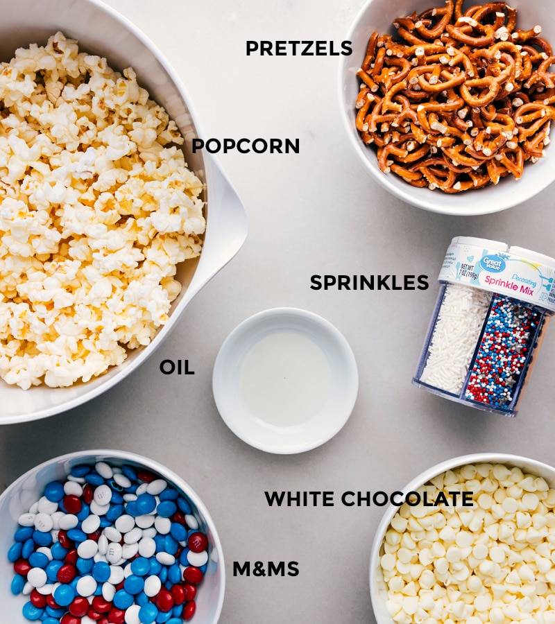 Ingredients In 4th Of July Popcorn All the ingredients for 4th of July Popcorn laid out, including popcorn, pretzels, sprinkles, white chocolate, and M&Ms, ready for easy assembly.