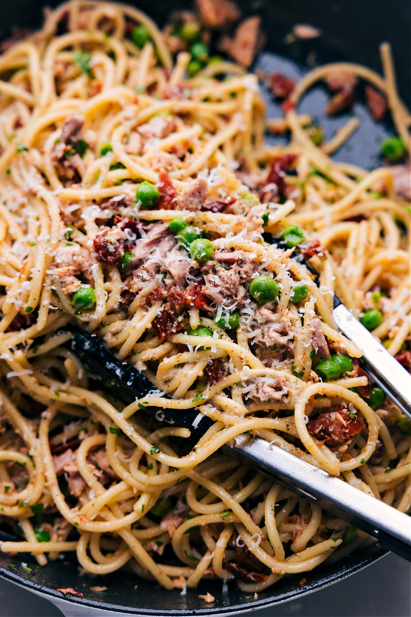 Serving Healthy Tuna Salad Pasta with Tongs Tongs scooping up a hearty portion of flavorful and healthy tuna salad pasta, garnished with freshly grated cheese, showcasing a blend of vibrant ingredients and fresh appeal.