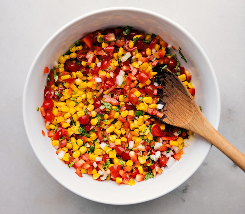 This Corn and Tomato Salad recipe features a vibrant blend of sweet, juicy corn and tomatoes balanced by the crunch of diced bell pepper and onion. The tangy lime vinaigrette, spiked with a hint of maple syrup and Dijon mustard, adds a burst of citrusy freshness that ties together everything beautifully! Recipe via ChelseasMessyApron.com #cornsalad #corntomatoes #corn #tomatoes #salad #lime #vinaigrette #summery #basil Overhead view of a bowl of Corn and Tomato Salad with a wooden spoon in the bowl.