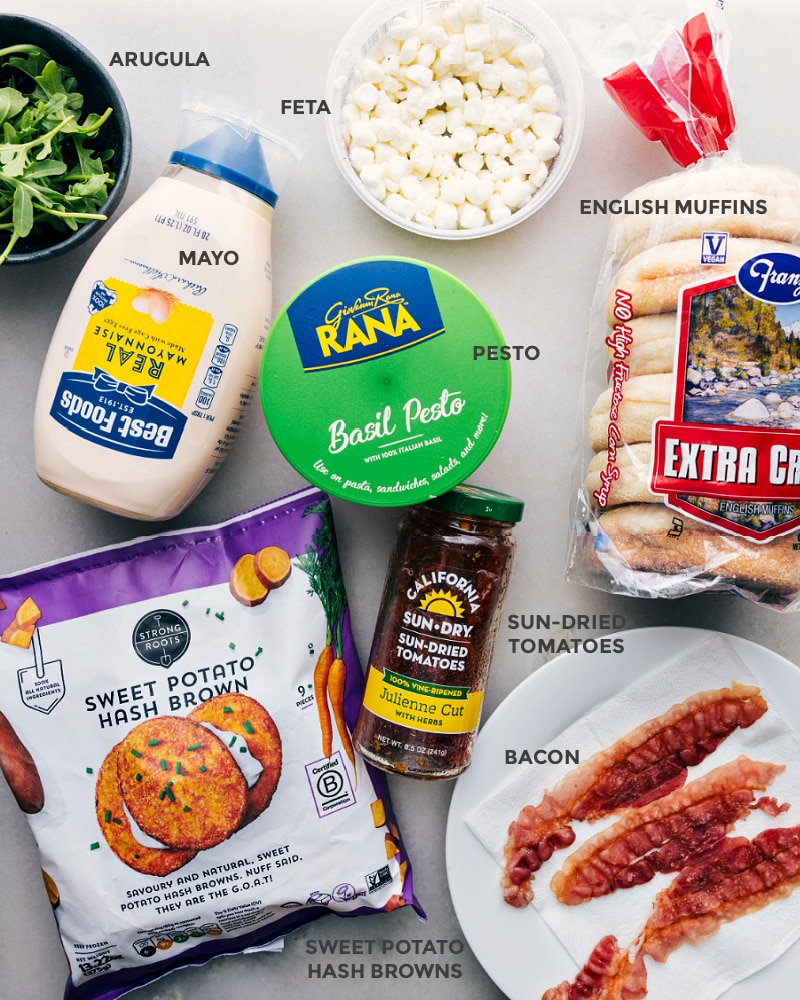 Ingredients In English Muffin Sandwich Ingredients prepped out for easy assembly including the arugula, feta, mayo, pesto, sun-dried tomatoes, bread, sweet potato hash browns, and bacon.