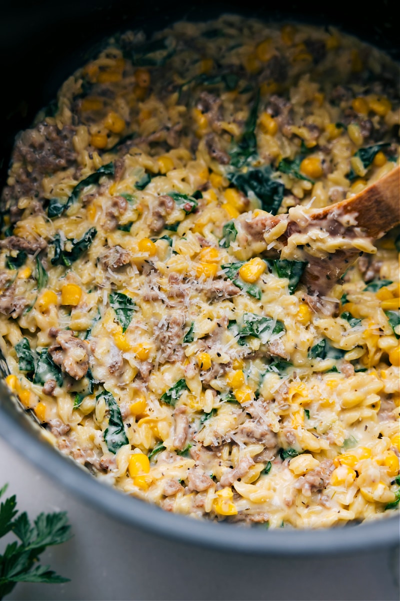 Sausage, Corn, & Spinach Orzo combines savory Italian sausage, sweet corn, and fresh spinach, all enveloped in a creamy, rich sauce jazzed up with Italian seasonings and Parmesan cheese. The dish is hearty, flavorful, and super comforting with a risotto-like creaminess. Recipe via chelseasmessyapron #dinner #quick #easy #simple #familyfriendly #kidfriendly #comfortfood #sausage #corn #spinach #orzo Up-close overhead image of the dish in a pot ready to be enjoyed