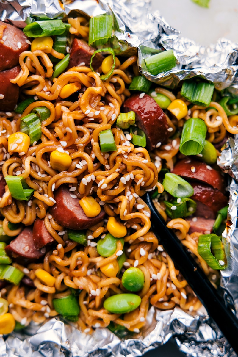 Our favorite Foil Pack Ramen Noodles recipe is an explosion of flavors, all conveniently packed into a single foil package. The dish unites the umami-rich, spicy, and slightly sweet notes of a quick homemade stir-fry sauce with the smokiness of sausage, freshness of veggies, and the comforting familiarity of ramen noodles.ย Recipe via chelseasmessyapron #dinner #quick #easy #simple #familyfriendly #kidfriendly #easycleanup #camping #campingfood #foilpack #ramennoodles Overhead image of the dish ready to be enjoyed
