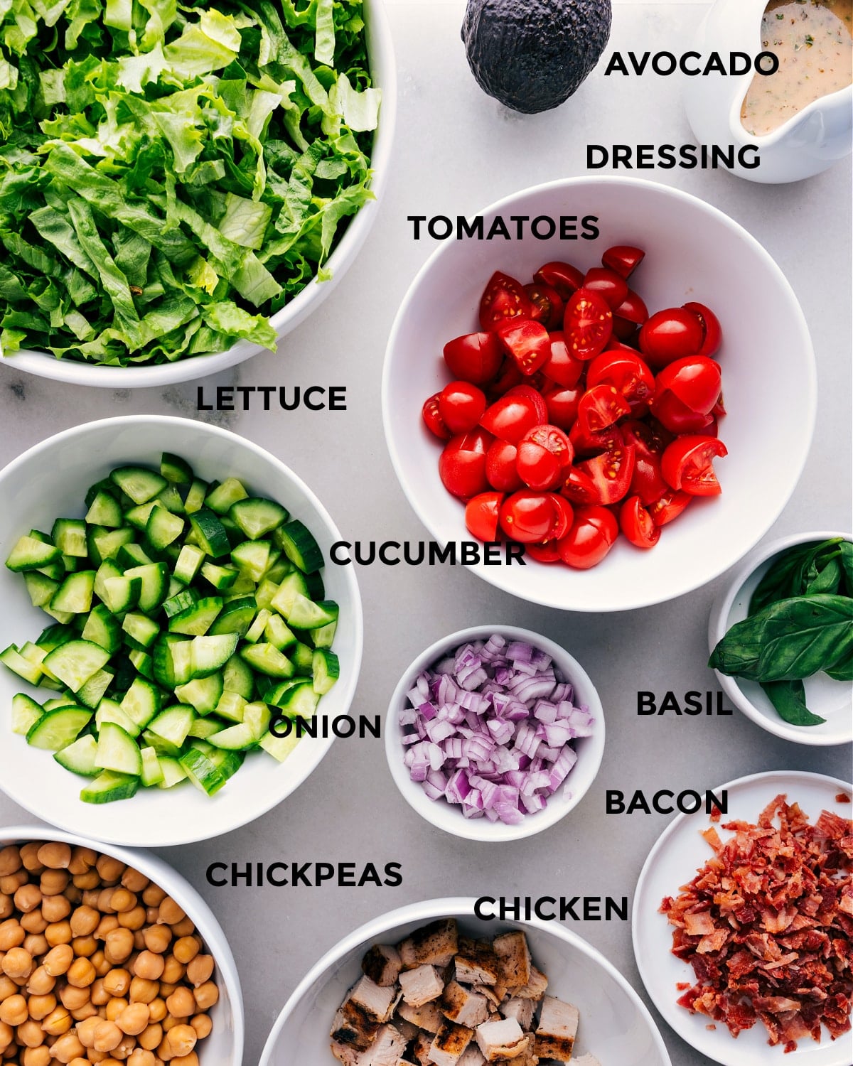 Ingredients In Chopped Chicken Salad This recipe has everything prepped for easy assembly: tomatoes, avocado, dressing, lettuce, cucumbers, onions, basil, bacon, chicken, and chickpeas.