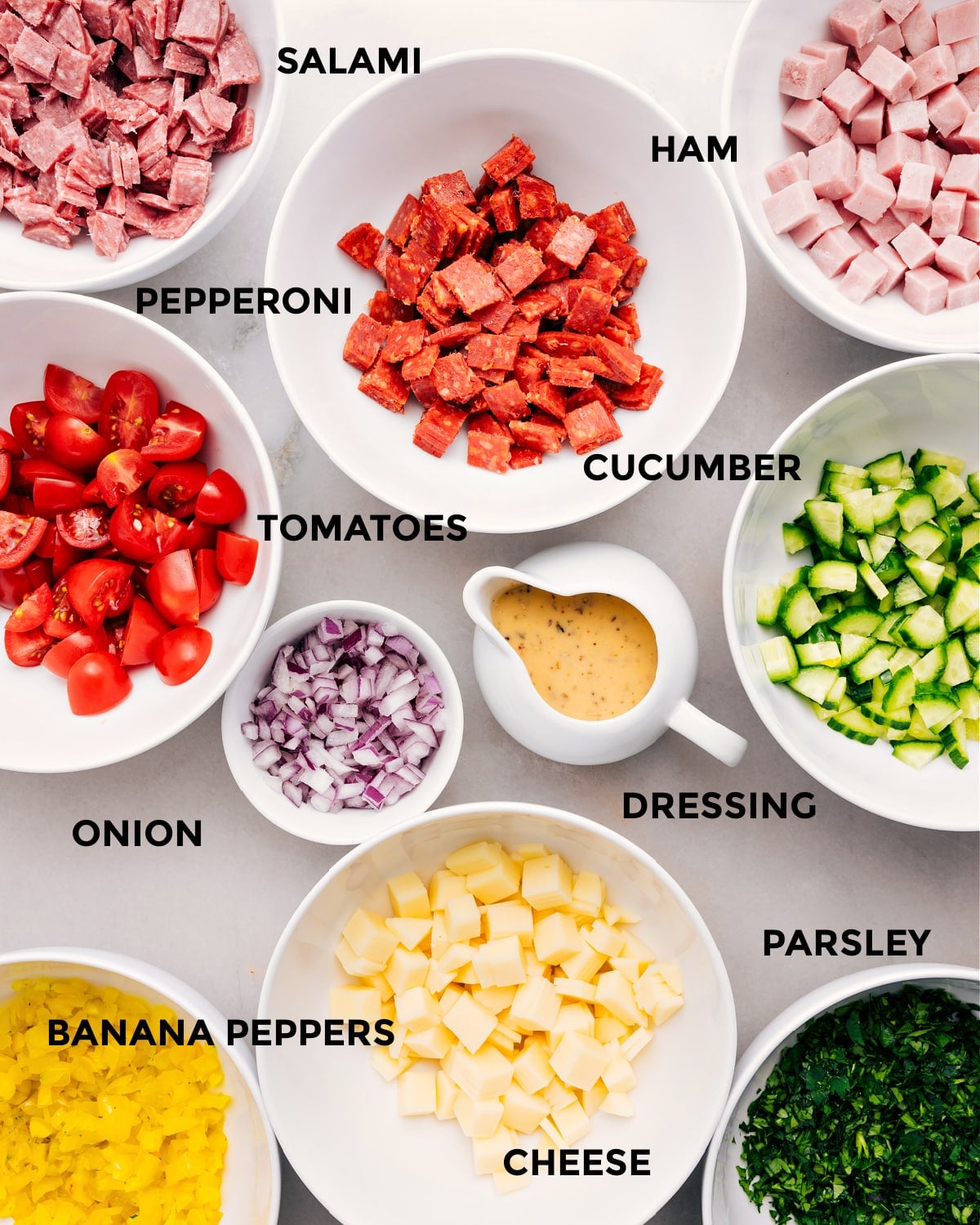 Ingredients In Italian Grinder Pasta Salad Recipe All the ingredients are prepped for easy assembly—meat, cucumber, tomatoes, onion, dressing, cheese, banana peppers, and parsley.
