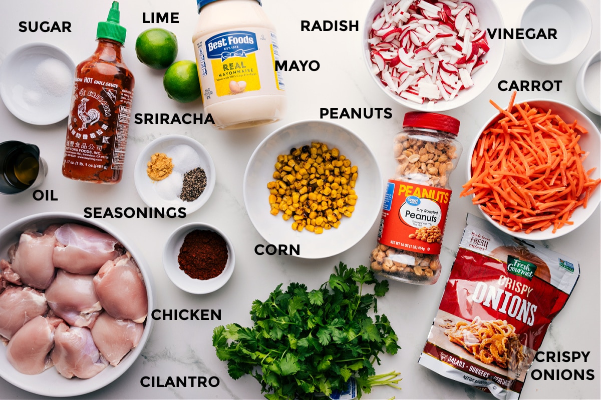 Ingredients In Asian Chicken Bowls All ingredients prepped for easy assembly: sugar, sriracha, lime, mayo, radish, vinegar, carrot, peanuts, corn, seasonings, oil, meat, cilantro, and crispy onions.